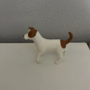 Schleich hund - En liten hundfigur i vitt och brunt. Figuren har en stående pose med svansen uppåt och bruna fläckar på kroppen och huvudet. Perfekt för samlare eller som dekoration.