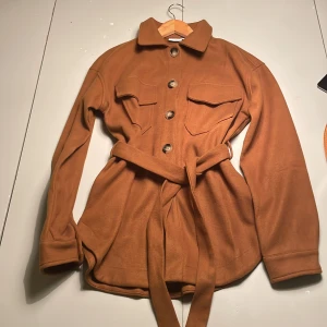 Oanvänd brun kappa med bälte - Säljer en snygg brun kappa med knappar och bälte i midjan. Jackan har långa ärmar och två bröstfickor med lock. Perfekt för höst och vår. Den har en klassisk krage och är tillverkad i ett mjukt material.