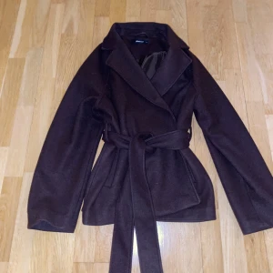 Brun kappa från Gina Tricot - Snygg brun kappa från Gina Tricot med knytskärp i midjan. Perfekt för höst och vår, med en klassisk design och långa ärmar. Tillverkad i ett mjukt material som ger en bekväm passform. Knappt använd typisk gina jacka i mörk brun💗