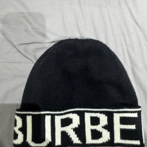 Svart mössa från Burberry - Snygg svart mössa från Burberry med vit text runt kanten. Perfekt för att hålla värmen med stil. Den är stickad och har en klassisk design som passar till det mesta.