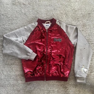 Röd och silver paljettjacka från Molo - Säljer en cool röd och silver paljettjacka från Molo med dragkedja framtill. Jackan har långa ärmar och en broderad text 'Unity' på bröstet.       Hubertha bomberjacka från MOLO är fylld med paljetter i röd med vita ärmar. Jackan har dragkedja och ribbkanter. Paljetterna kan borstas upp så att det blir ännu mer spel i dem. Ordinarie pris :799kr 	 Perfekt för att sticka ut med sin glittriga design!