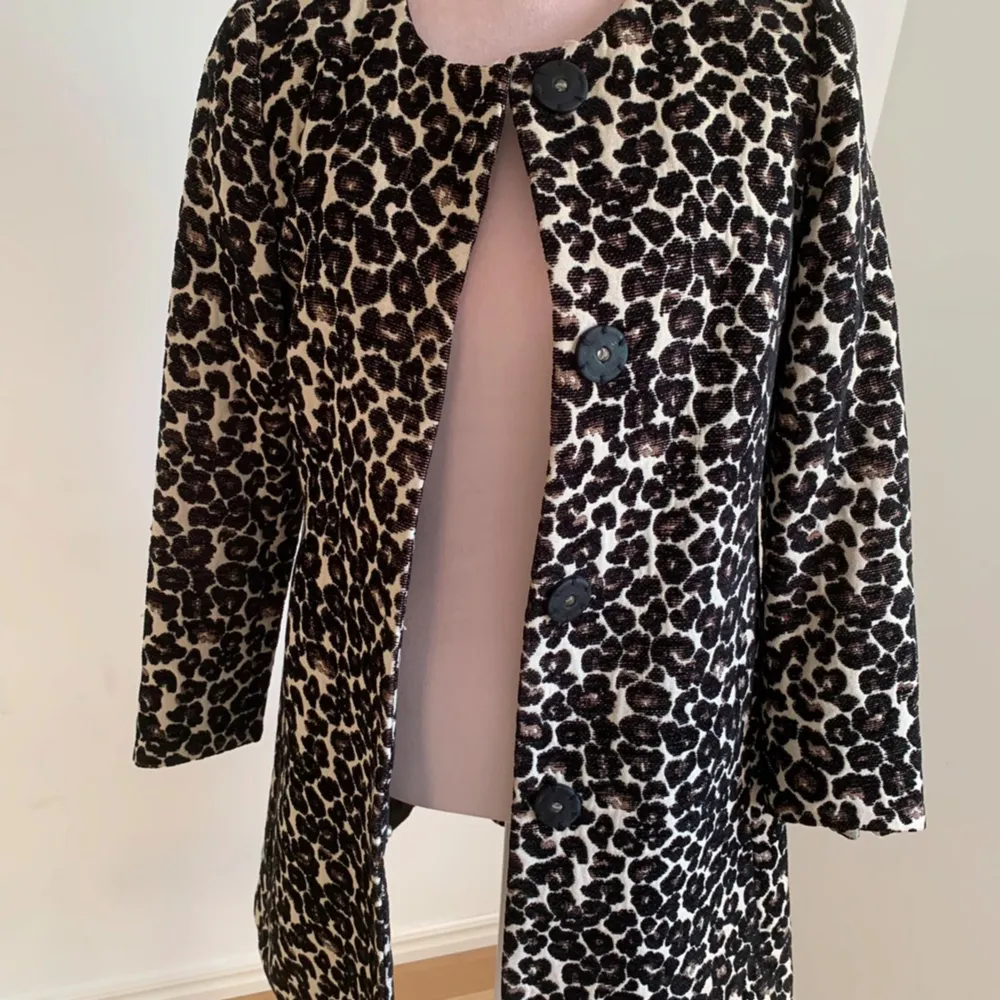 Snygg leopardmönstrad kappa med stora svarta knappar framtill. Den har en klassisk passform och är perfekt för att ge en trendig touch till din outfit. Kappan har långa ärmar och en rund halsringning.. Takit.