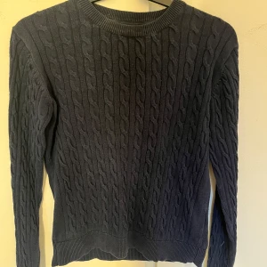 Mörkblå kabelstickad tröja - Säljer en stilren mörkblå kabelstickad tröja med rund halsringning. Tröjan har långa ärmar och ribbade muddar vid ärmslut och nederkant. Perfekt för kyliga dagar.