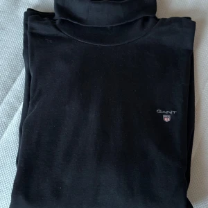 Svart turtleneck från GANT - Snygg svart turtleneck från GANT med diskret logga på bröstet. Perfekt för en stilren look. Tröjan har långa ärmar och en klassisk turtleneck krage. 