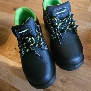Svarta arbetsskor från Bluewear - Säljer ett par robusta svarta arbetsskor från Bluewear med grönt foder och snörning. Skorna har en slitstark sula och är perfekta för arbete där extra skydd behövs. De har en klassisk design med kontrasterande sömmar och en bekväm passform.
