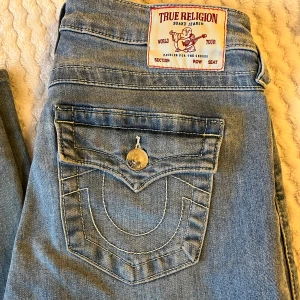 Blå jeans från True Religion - Säljer ett par snygga blå jeans från True Religion med ikoniska bakfickor och knappdetaljer. Jeansen har låg midja och bootcut! På sista bilden ser man lite skada på insidan av innerbenet, knappt synlig men fråga om du har fler funderingar!💕säljer då de var lite för stora för mig och har köpt nya!