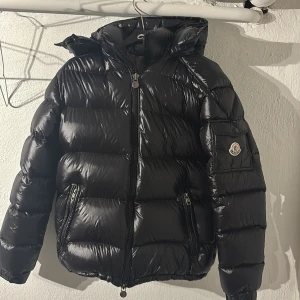 ( ÄKTA )Moncler Maya  - Säljer en snygg svart dunjacka från Moncler. Jackan har en glansig finish och är utrustad med en dragkedja framtill. Den har en huva och långa ärmar, perfekt för kyligare dagar. Insidan har ett unikt serietidningsmönster som ger en cool touch. Jag hade kunnat sänka priset 