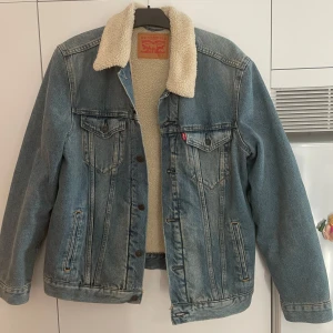 Jeansjacka med teddyfoder från Levi's - Säljer en snygg jeansjacka från Levi's i storlek L. Den har ett mysigt teddyfoder som gör den perfekt för höst och vinter. Jackan har en klassisk blå jeansfärg och är långärmad med knappar framtill. 