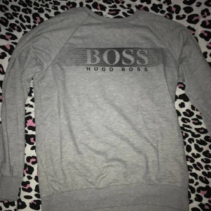 Grå tröja från Hugo Boss - Säljer en stilren och bekväm grå tröja från Hugo Boss. Den har ett stort tryck med märkesnamnet på framsidan och är tillverkad i ett mjukt material. Perfekt för en avslappnad look!