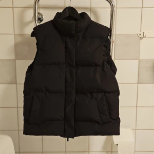 Svart dunväst i återvunnen polyester - Dunväst i 100% återvunnen poly från Knowledge Cotton. Vindtät och har en andningsförmåga på 5,000g/m2 24h samt är vattentät upp till 5,000mm. Håller dej både varm och torr. Den stängs med dragkedja sam magnetknapp. Inneficka.