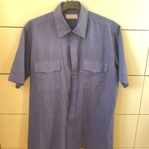 Blå kortärmad skjorta från Pilot - Säljer en snygg blå kortärmad skjorta från Pilot. Den är i storlek M.  Den är både bekväm och lättskött. Skjortan har två bröstfickor med lock och knappar framtill. Perfekt för sommardagar eller avslappnade tillfällen!😃