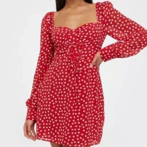 Röd blommig klänning från ADOORE  - Superfin röd klänning med vita blommor från Adoore. Den har en söt knytning framtill och långa ärmar. Perfekt för sommarens fester eller en mysig vårdag. Klänningen är kort och har en smickrande passform. Oanvänd.