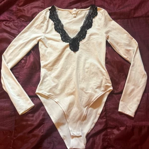 Vit långärmad body med spetsdetaljer - Säljer en superfin vit body med långa ärmar och snygg svart spets runt den v-ringade halsen. Perfekt för en stilren look, både till vardags och fest. Materialet känns mjukt och skönt mot huden. Passar perfekt till höst- och våroutfits!