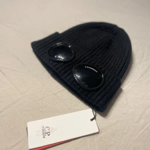 Svart C.P. Company mössa - Snygg svart ribbad mössa från C.P. Company Perfekt för att hålla sig varm och stilren under kalla dagar. Mössan är helt ny och har en klassisk passform som passar de flesta.