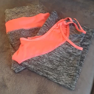 Grå och neonorange träningsset - Säljer ett snyggt träningsset i grått och neonrosa. Setet består av en topp och byxor, perfekt för gymmet eller löprundan. Materialet är stretchigt och bekvämt, vilket ger en bra passform. Toppen har en sportig design med brottarrygg. Säljes tillsammans.