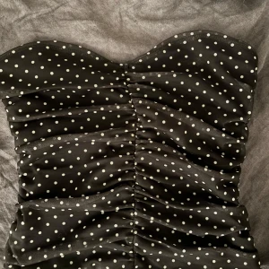 Svart prickig tubtopp - Snygg svart tubtopp med vita prickar. Toppen har en rynkad design som ger en cool look. Perfekt för sommarens fester eller en utekväll. Den är axelbandslös och har en tight passform som framhäver figuren. 