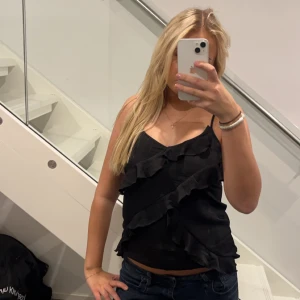 Svart volangtopp från B Angel - Snygg svart topp från B Angel med volangdetaljer. Den är ärmlös och har smala axelband, perfekt för en utekväll eller fest. Toppen är i bra skick och ger en elegant touch till vilken outfit som helst.