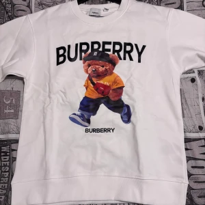 Vit sweatshirt från Burberry - Säljer en superfin vit sweatshirt från Burberry med ett coolt nalletryck på framsidan. Nallen har en orange tröja och en svart keps, vilket ger en lekfull touch. Perfekt för en avslappnad stil och passar till både jeans och joggers. Tröjan är i bra skick och redo för nya äventyr!
