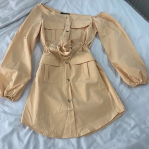 Beige klänning med knappar - Säljer en superfin beige klänning med knappar en och långa ärmar. Den har en snygg knytning i midjan och praktiska fickor på bröstet. Perfekt för både vardag och fest!