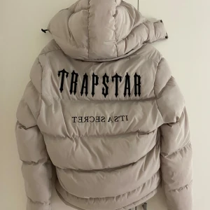 Beige pufferjacka från Trapstar - Säljer en snygg beige pufferjacka från Trapstar i storlek S. Jackan har en cool logga på ryggen och en dragkedja framtill. Perfekt för kalla dagar med sin varma vaddering och huva. Den är i mycket bra skick och redo för nya äventyr!