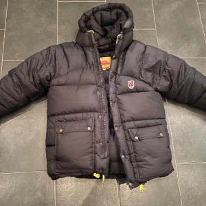 Svart dunjacka från Fjällräven - Säljer en svart dunjacka från Fjällräven Expedition down i mycket bra skick. Jackan har en quiltad design med dragkedja och knappar framtill. Perfekt för kalla vinterdagar med sin varma och bekväma passform. Den har långa ärmar och praktiska fickor med knappar. En klassisk och stilren jacka för alla tillfällen.