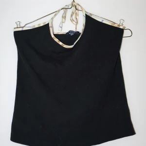 Svart halterneck topp från Burberry - Snygg svart halterneck topp från Burberry i bomull. Den har ett klassiskt rutigt mönster i beige och lila runt halsen. Perfekt för en stilren och elegant look. Passar både till vardags och fest!