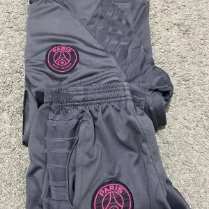 Grå PSG shorts med rosa detaljer - Säljer ett par grå PSG shorts från Nike med snygga rosa detaljer och logga. De har en elastisk midja och dragkedjefickor. Perfekta för fotbollsträningen eller bara för att chilla i. Materialet känns skönt och andas bra. Passar perfekt för sommarens aktiviteter! Priset är ej hugget i sten!