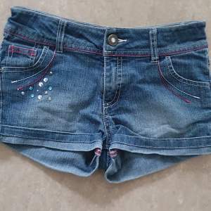 Snygga blå jeansshorts från Mywear Young i storlek 134. De har coola strassdetaljer på framsidan och kontrastsömmar i rosa. Perfekta för sommardagar och avslappnade tillfällen. Shortsen har en knapp och dragkedja framtill samt fickor både fram och bak.