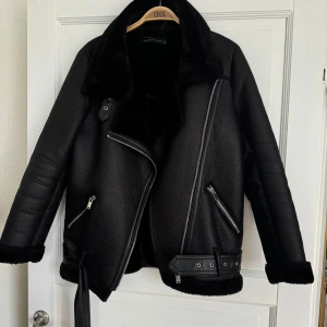 Svart Zara jacka med pälsdetaljer - Säljer en snygg svart zara jacka med pälsdetaljer på kragen och ärmsluten. Jackan har en cool asymmetrisk dragkedja och bälte i midjan för extra stil. Perfekt för höst och vinter, håller dig varm och trendig. Passar både till vardags och fest! Som ny!! 