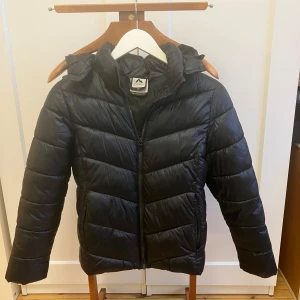 Svart pufferjacka från McKinley - Säljer en svart pufferjacka från McKinley i mycket bra skick. Jackan har en dragkedja framtill och en praktisk huva. Perfekt för kalla dagar med sin vadderade design som håller värmen. Passar både till vardags och utomhusaktiviteter.
