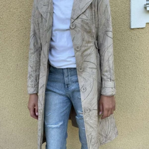 Beige trenchcoat med blommönster - Säljer en elegant beige trenchcoat med subtilt blommönster. Jackan har långa ärmar och knappar framtill. Den är perfekt för höst och vår, och ger en stilren look. Passar både till vardags och festliga tillfällen. Storlek 34, från Green house 