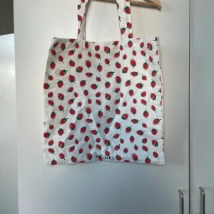 Vit shoppingväska med jordgubbsmönster från Monki - Superfin vit shoppingväska från Monki med ett gulligt jordgubbsmönster. Perfekt för sommarens äventyr eller en tur på stan. Väskan har långa handtag som gör den enkel att bära över axeln. Ett måste för alla som älskar fruktiga detaljer! 🍓