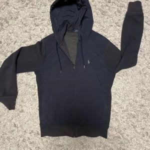 Mörkblå hoodie från Ralph Lauren - Säljer en snygg mörkblå hoodie från Ralph Lauren i mycket bra skick. Den har en dragkedja framtill och en broderad logga på bröstet. Perfekt för höst och vår och vinter, med långa ärmar och en skön huva. Passar både till vardags och lite mer avslappnade tillfällen.