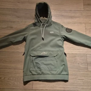 Grön jacka från Dope - Säljer en snygg grön jacka från Dope. Den har en stor ficka framtill med ett coolt bergsmotiv och en justerbar huva med dragsko. Perfekt för kyliga dagar och har en avslappnad passform. Märkesdetaljer på bröstet och ärmen ger en extra touch. Nästan aldrig använd därav priset ❤️