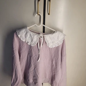 Ljuslila tröja med vit krage från Kallinera - Säljer en söt ljuslila tröja från Kallinera med en stor vit krage som har volangdetaljer. Perfekt för en chic och vintage-inspirerad look. Tröjan har långa ärmar och är i bra skick. Passar perfekt till både vardag och fest!
