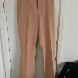 Säljer ett par stilrena beige/aprikos kostymbyxor från Bik Bok i storlek 38. Perfekta för både kontoret och festligare tillfällen. Byxorna har en klassisk rak och lite bootcut i slutet och är tillverkade i ett bekvämt material. De är i bra skick och redo för en ny garderob!