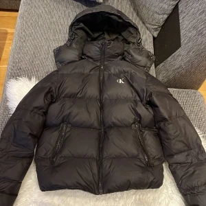 Svart pufferjacka från Calvin Klein Jeans - Säljer en svart pufferjacka från Calvin Klein Jeans i storlek XS. Jackan har en mysig huva och dragkedja framtill. Perfekt för kalla dagar! Den är i bra skick med en liten reva på ärmen. Passar både höst och vinter. Lite puffer försvann när jag skulle tvätta den men den är fortfarande varm och jätte bra mot de kyliga värdret. Nypris 2600