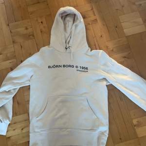 Säljer en stilren vit hoodie från Björn Borg med tryck '1965 Stockholm' på bröstet. Den har en klassisk känguruficka och justerbar huva. Perfekt för en avslappnad stil eller träning. Passar både höst och vår! Den har någon missfärgning på framsidan som syns väldigt lite. 