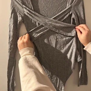 Grå omlottopp - Säljer en snygg grå omlottopp med långa ärmar. Perfekt för både vardag och fest. Toppen har en stilren design med omlottdetaljer framtill som ger en smickrande passform. Materialet är mjukt och stretchigt för extra komfort.