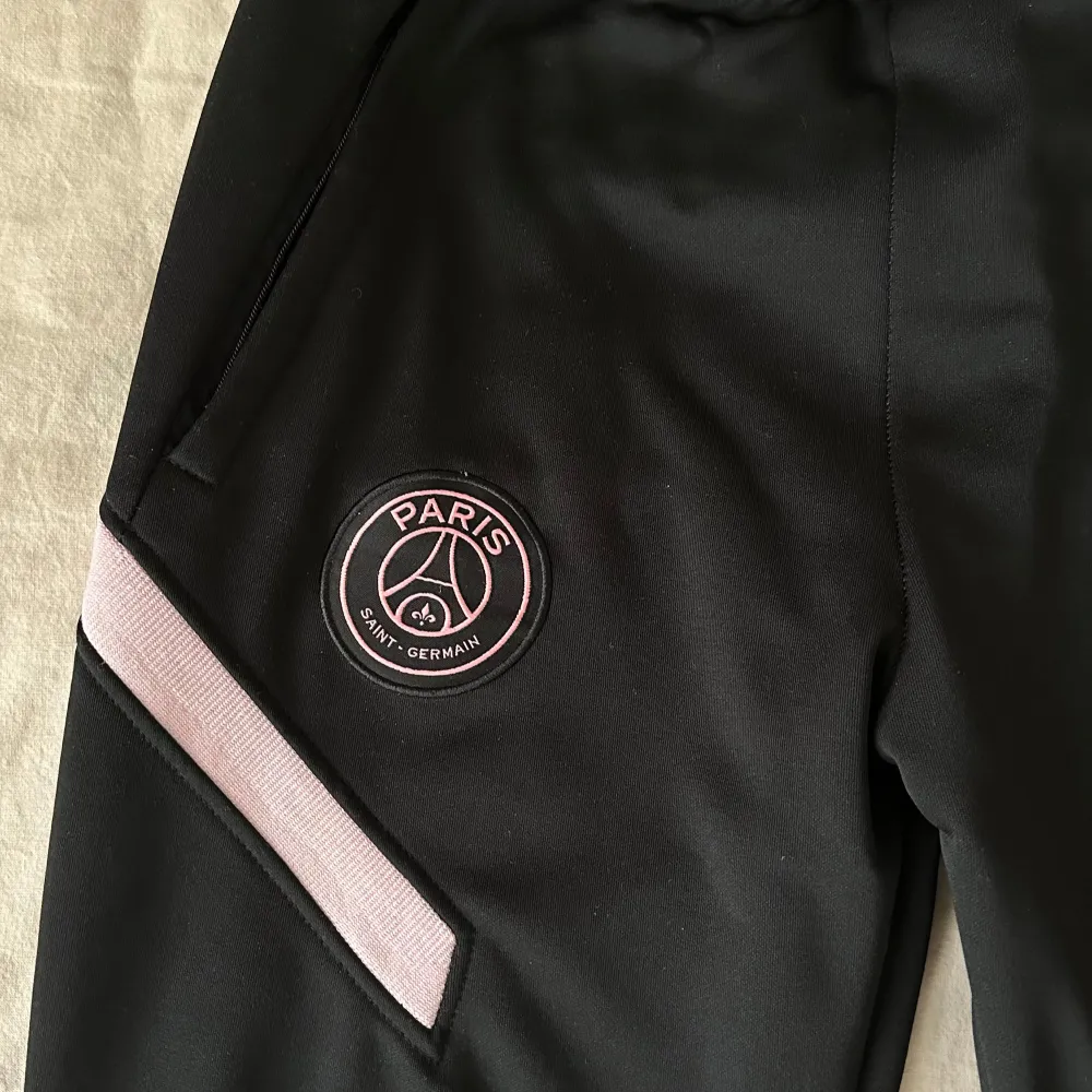 Säljer ett par svarta träningsbyxor från Nike- PSG. Byxorna har rosa detaljer och PSG-logga på ena benet. Perfekta för träning eller vardagsbruk. De har en elastisk midja och är gjorda i ett bekvämt material. Använd fåtal gånger, fortf mkt bra skick!!. Jeans & Byxor.