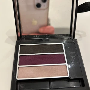 Dior Twist Trio Compact Eyeshadows - Säljer en elegant ögonskuggspalett från Dior med tre nyanser: mörkbrun, djup lila och ljus beige. Perfekt för både vardag och fest! Kommer i en snygg svart förpackning med ett unikt snörningsdetalj på baksidan. Paletten är kompakt och lätt att ta med sig.