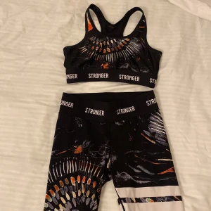 Svart träningsset från Stronger - Snyggt träningsset från Stronger i svart med coolt mönster i orange och vitt. Setet består av en sport-bh och tights. Materialet är stretchigt och bekvämt, vilket ger en tight passform. Både bh och tights har elastiska band med Stronger-loggan. Storlek S på båda! Hör gärna av er vid intresse! Köpte båda för totalt 1300 kr. Säljer för 400🥰