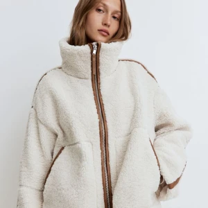 Vit teddyjacka med dragkedja - Supermysig vit teddyjacka med hög krage och dragkedja framtill. Perfekt för kyliga dagar och ger en stilren look med sina bruna detaljer. Jackan har en loose passform och är långärmad, vilket gör den idealisk för både höst och vinter. Aldrig använd med prislapp kvar!