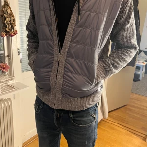 Grå cardigan stickade ärmar - Säljer en grå cardigan med stickade ärmar och krage. Perfekt för höst och vår, med dragkedja och ribbstickade muddar. Okänt märke.