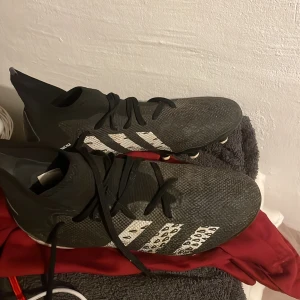 Svarta fotbollsskor med leopardmönster från addidas predator  - Säljer ett par svarta fotbollsskor i syntetmaterial med coola leopardmönstrade detaljer. Skorna har snörning och är i mycket bra skick. Perfekta för fotbollsträningen eller matchen. Storlek är inte angiven, men de ser ut att passa en normal passform.