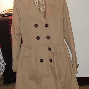 Beige trenchcoat med leopardmönstrat foder - Säljer en snygg beige trenchcoat med leopardmönstrat foder. Den har knappar framtill och ett bälte i midjan för en perfekt passform. Jackan är långärmad och passar perfekt för höst och vår. Perfekt för att ge din outfit en klassisk touch!