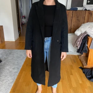 Svart lång kappa - Säljer en stilren svart lång kappa i bra skick. Den har en klassisk design med knappar framtill och en elegant passform. Perfekt för höst och vinter, och funkar både till vardags och fest. Kappan är långärmad och har en snygg krage.