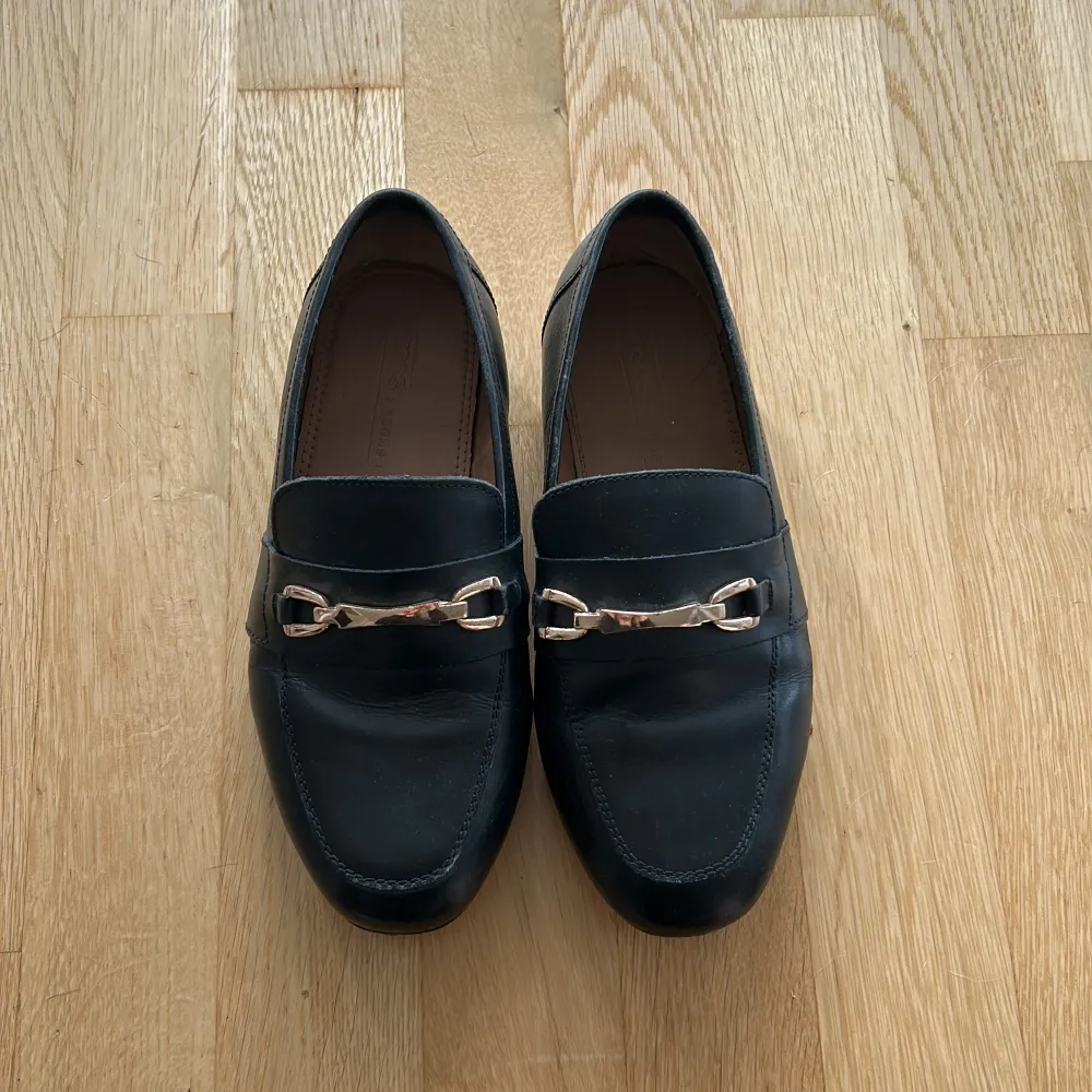 Säljer ett par stilrena svarta loafers i skinn med ett snyggt metallspänne framtill. Perfekta för både vardag och fest. De har en klassisk design och är superbekväma att ha på sig. Passar till nästan alla outfits!. Skor.