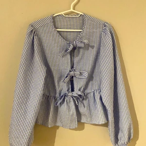 Blå och vit rutig blus med knytband - Säljer en superfin blå och vit rutig blus med knytband framtill. Den har långa ärmar och en söt volangdetalj vid midjan. Perfekt för vår och sommar! 💙