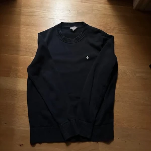 Mörkblå  tröja från Morris - Säljer en stilren svart tröja från Morris med rund halsringning och ett litet broderat emblem på bröstet. Tröjan har långa ärmar och ribbade muddar vid ärmslut och nederkant. Perfekt för en klassisk look.